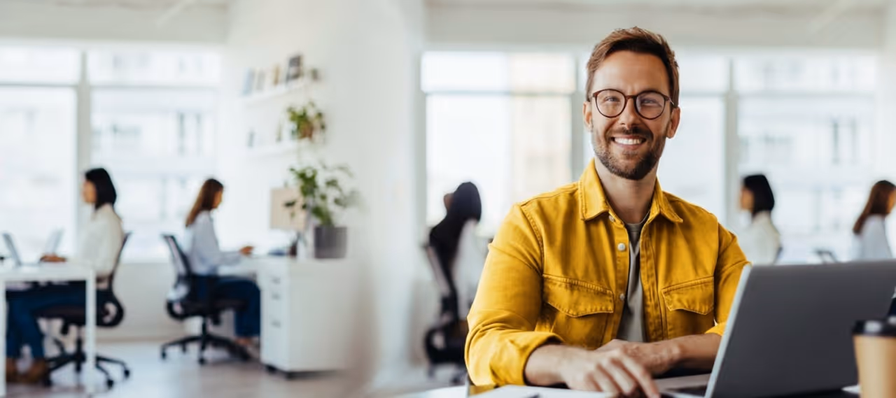 Smiling man with glasses and yellow jacket working on a laptop in a modern office with coworkers.
