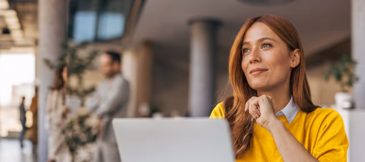 Woman in yellow sweater thoughtfully looking away while seated with a laptop in a modern office.
