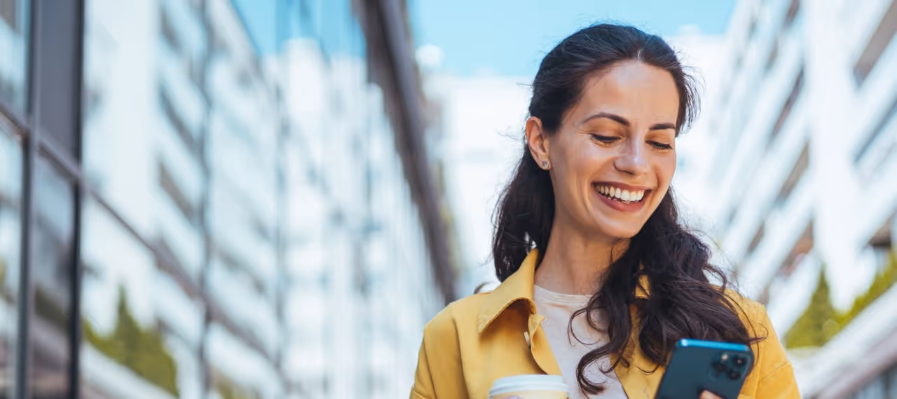 Smiling woman in yellow jacket holding coffee cup and looking at smartphone outside between buildings.