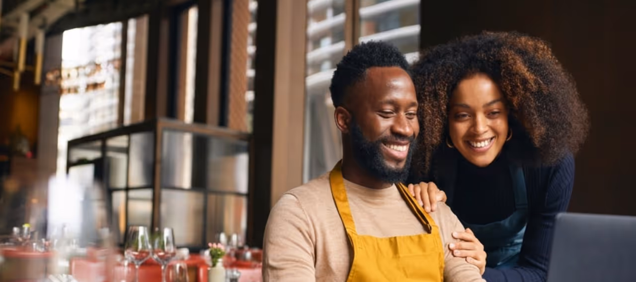 Smiling man in yellow apron and woman stand behind him in a warmly lit restaurant interior.