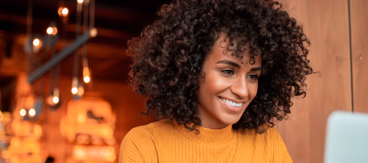 Smiling woman with curly hair wearing a mustard sweater in a warmly lit indoor setting.