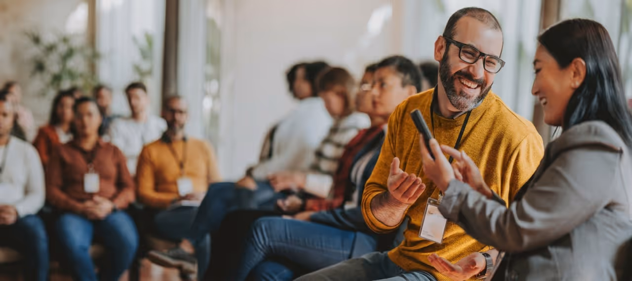 Man in glasses and yellow sweater smiling at woman showing something on smartphone during group meeting.
