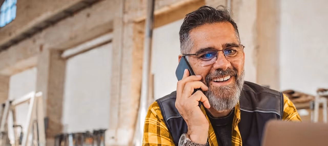 Smiling man with beard wearing glasses and a yellow plaid shirt talking on a phone in an indoor setting.