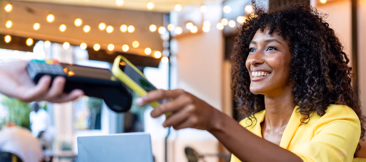 Smiling woman in yellow jacket holding phone for contactless payment at a café with string lights.