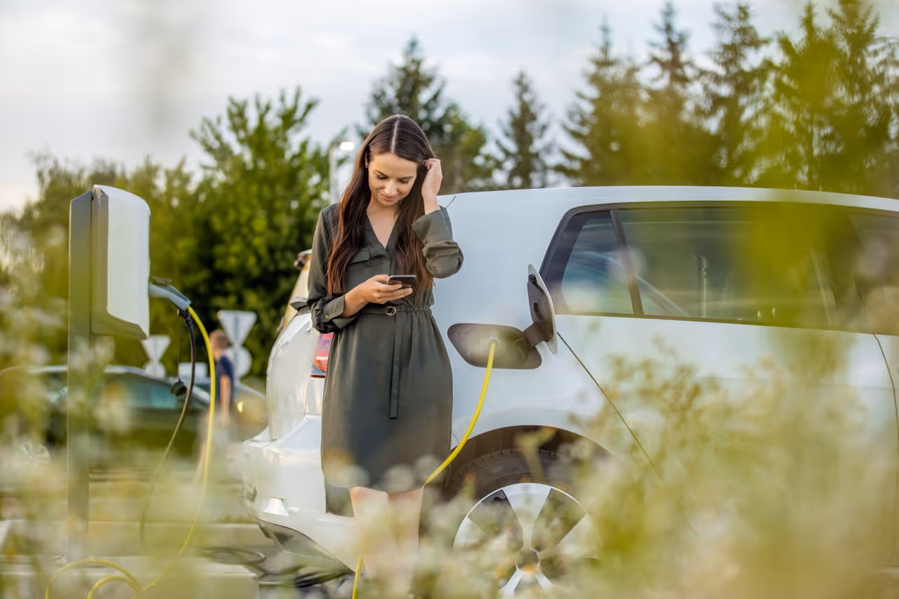 Woman in green dress using smartphone near white electric car plugged into a charging station.