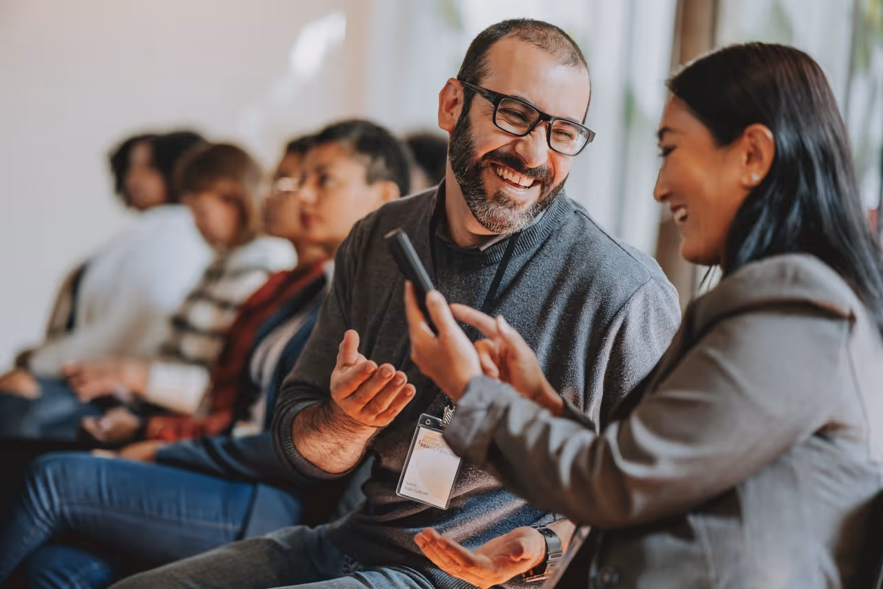Man with glasses smiling and talking to woman showing phone, others seated in a row behind them.