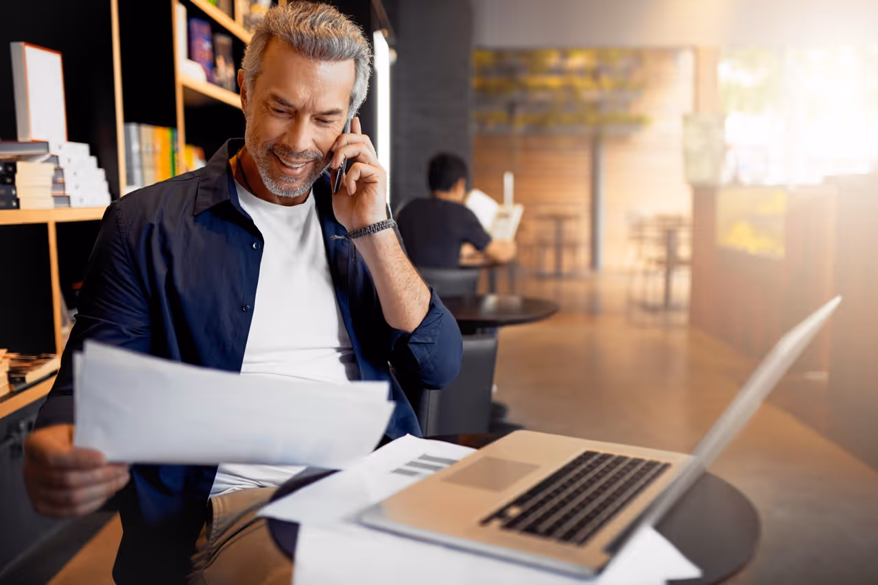 Smiling man talks on phone reviewing documents with laptop in bright cafe setting.