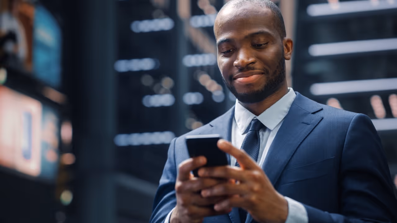 Smiling man in blue suit looking at his smartphone with blurred city lights in the background.