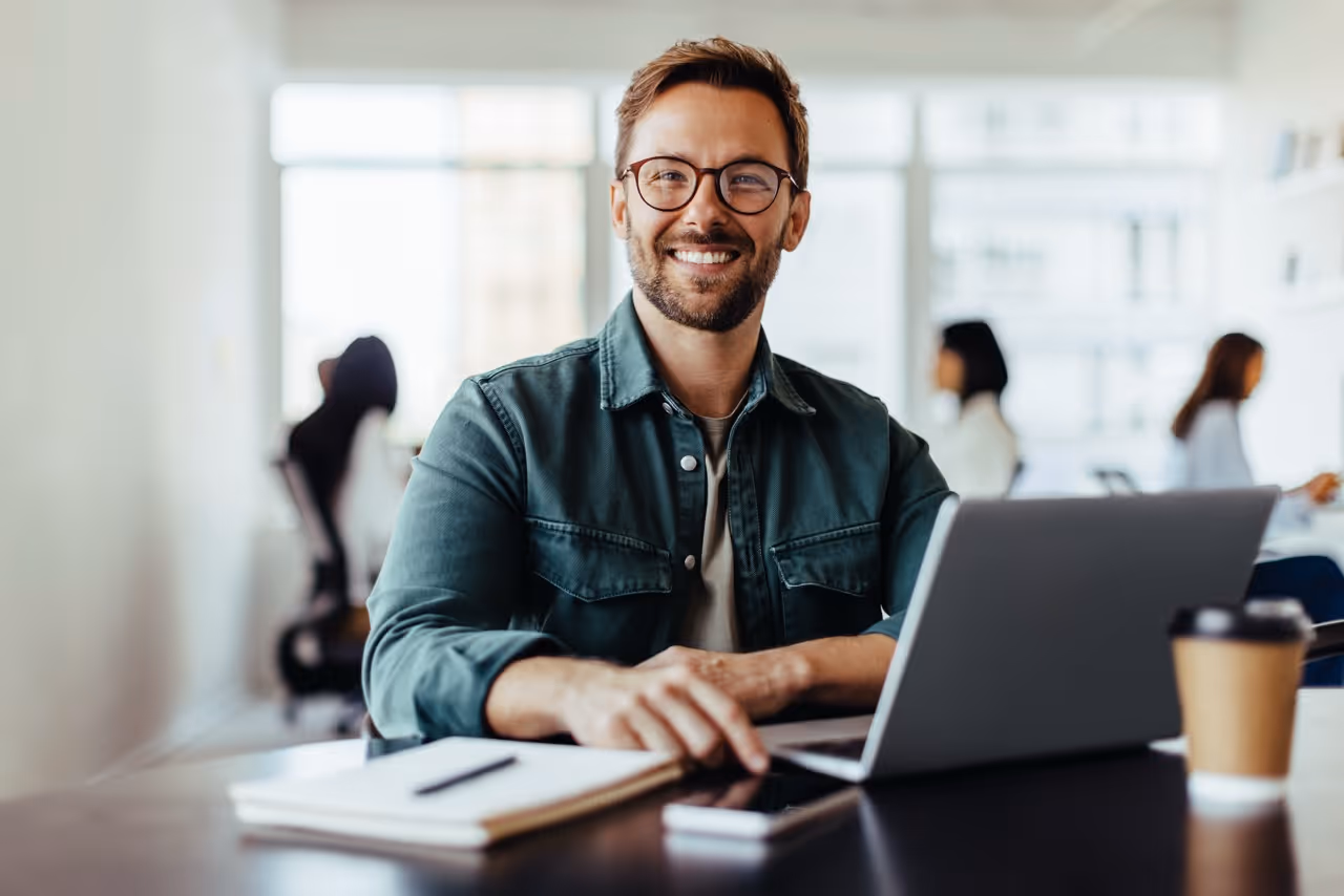 Smiling man wearing glasses seated at desk with laptop, notebook, smartphone, and coffee in office.