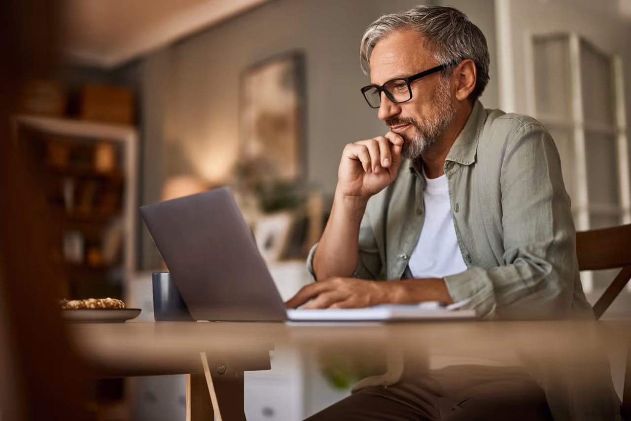 Mature man wearing glasses focused on laptop screen while seated at a dining table in a cozy room.
