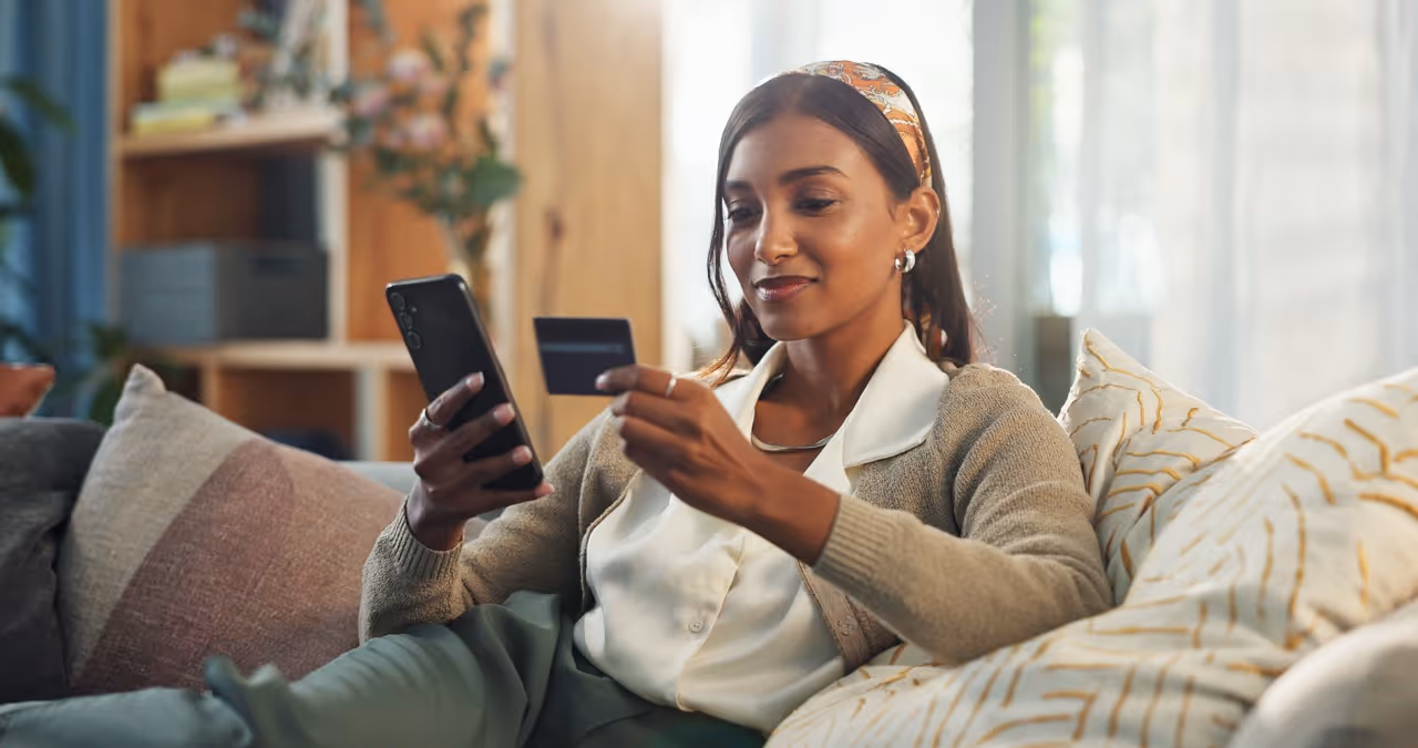 Woman sitting on couch holding phone in one hand and credit card in the other, shopping online.
