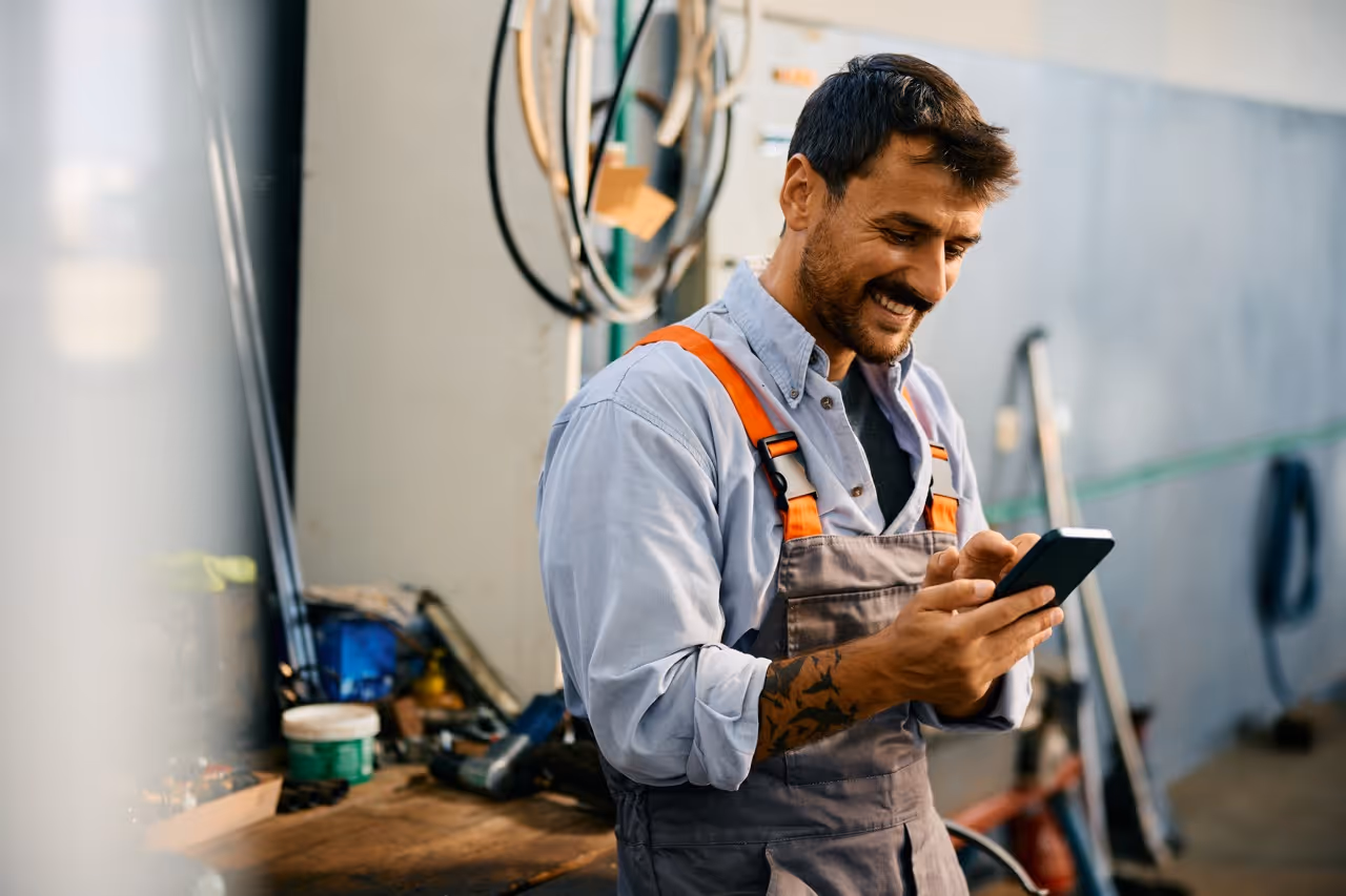 Mechanic in work overalls smiling while using a smartphone in a workshop.