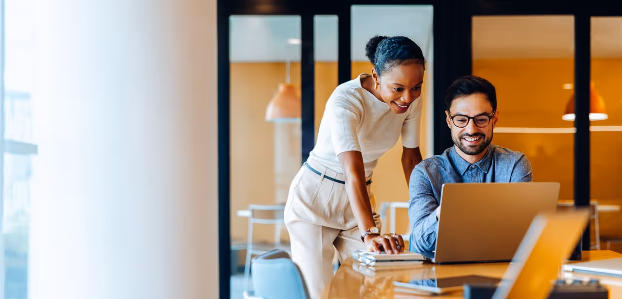 Two coworkers smiling and looking at a laptop screen in a modern office space.