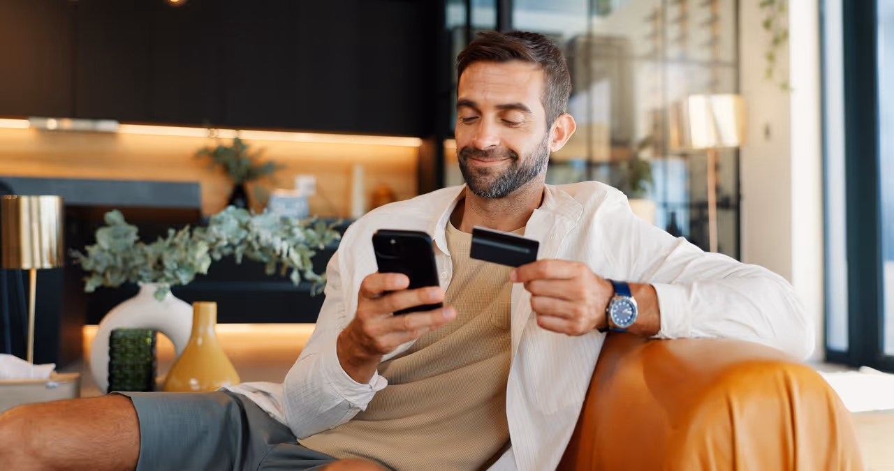 Man sitting on a couch using a smartphone and holding a credit card, smiling slightly.