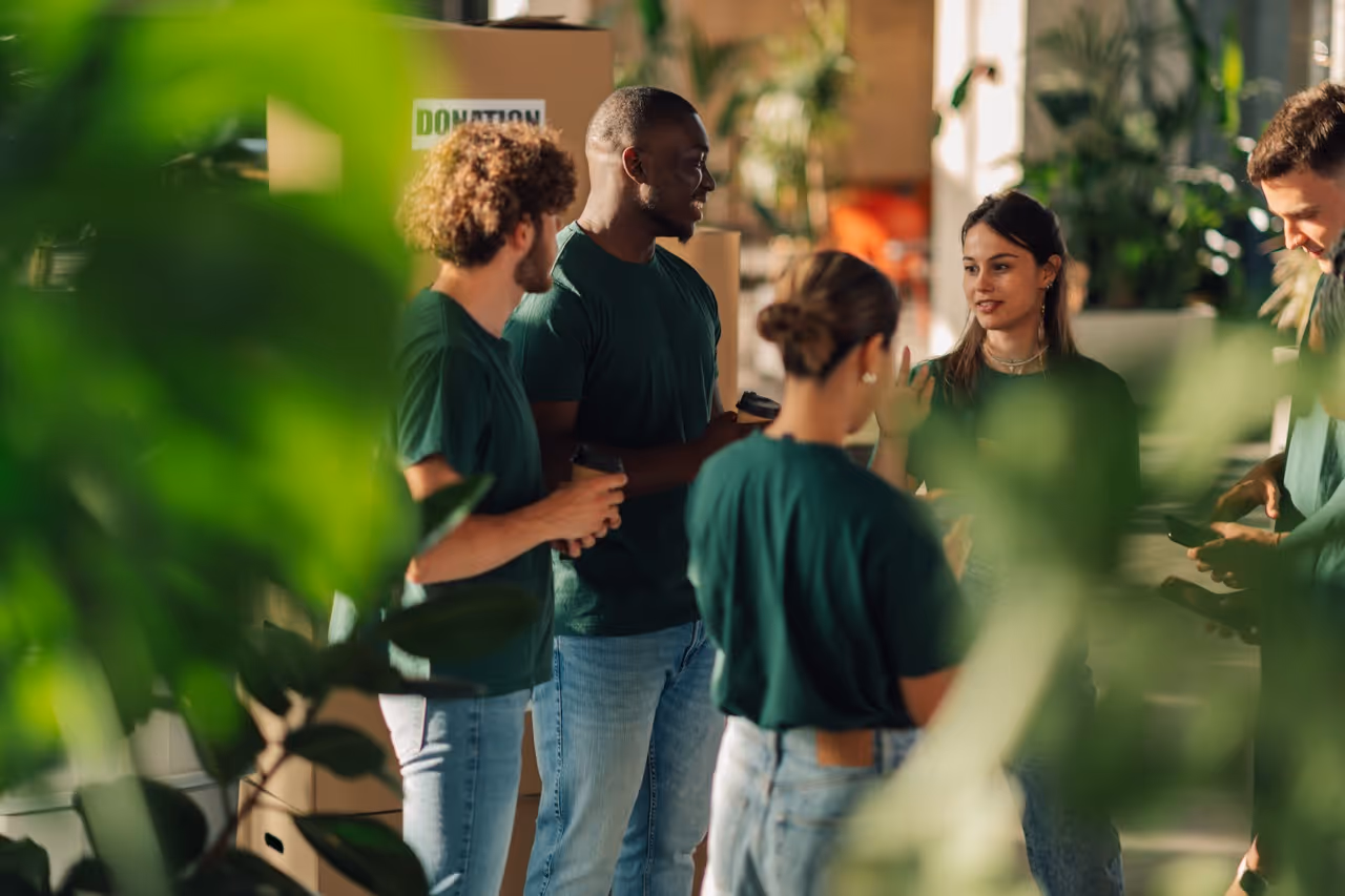 Diverse group of young adults in green shirts chatting near donation boxes surrounded by plants.