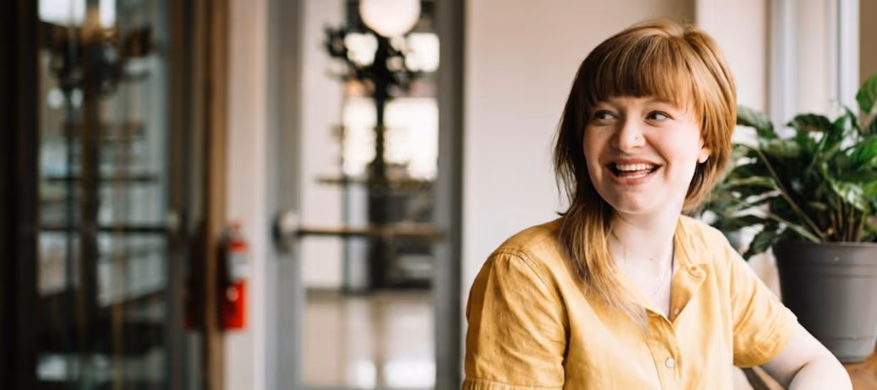 Smiling woman with short red hair wearing a yellow shirt sitting indoors near a plant.