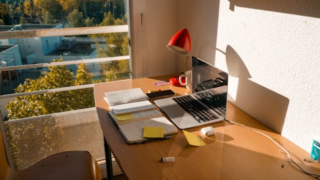 Workspace with laptop, books, sticky notes, red lamp, and coffee mug on wooden desk near window.