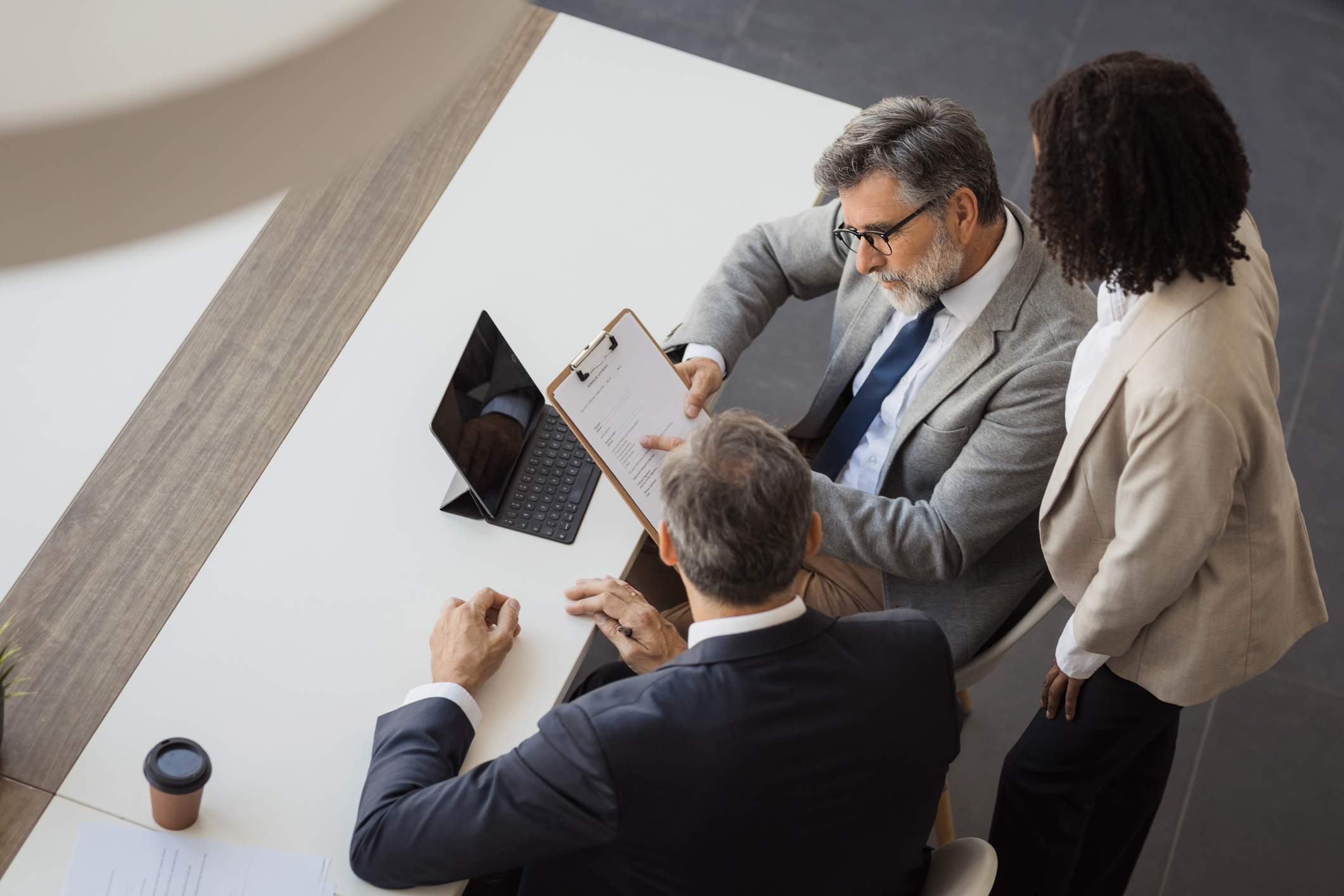 Three business professionals reviewing a document on a clipboard at a white table with a tablet and coffee cup.