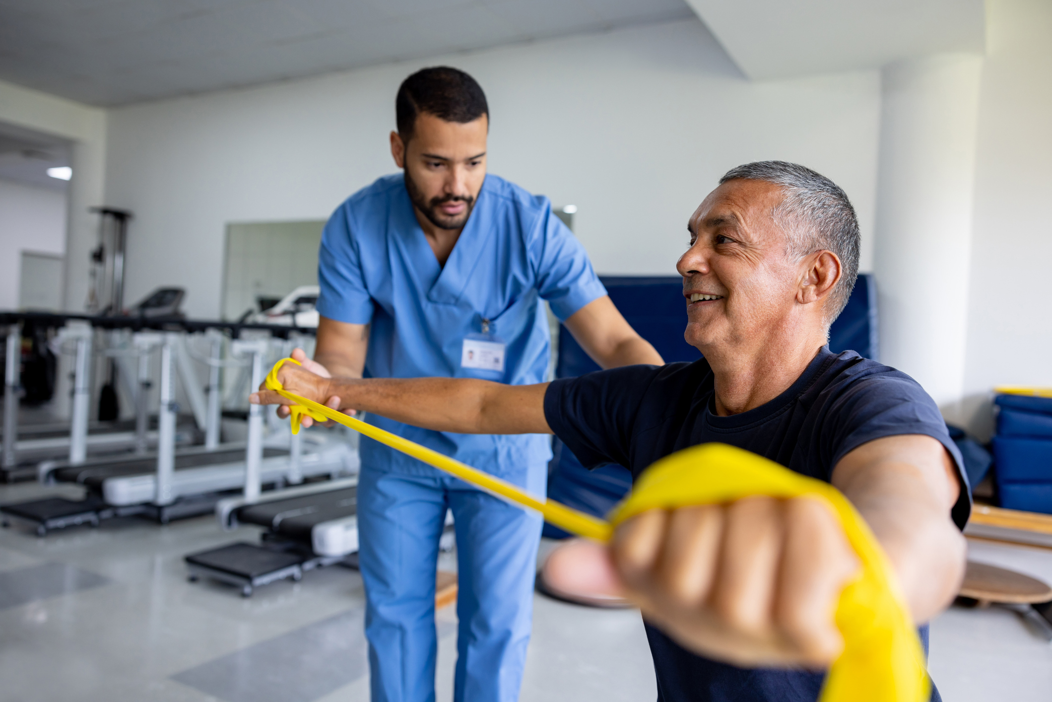 Older man exercises with yellow resistance band guided by a therapist in a rehab gym.