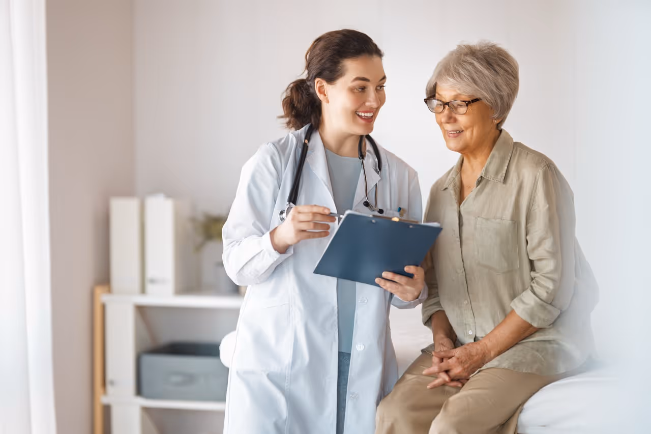 Female doctor with stethoscope discussing notes on clipboard with elderly woman in clinic.