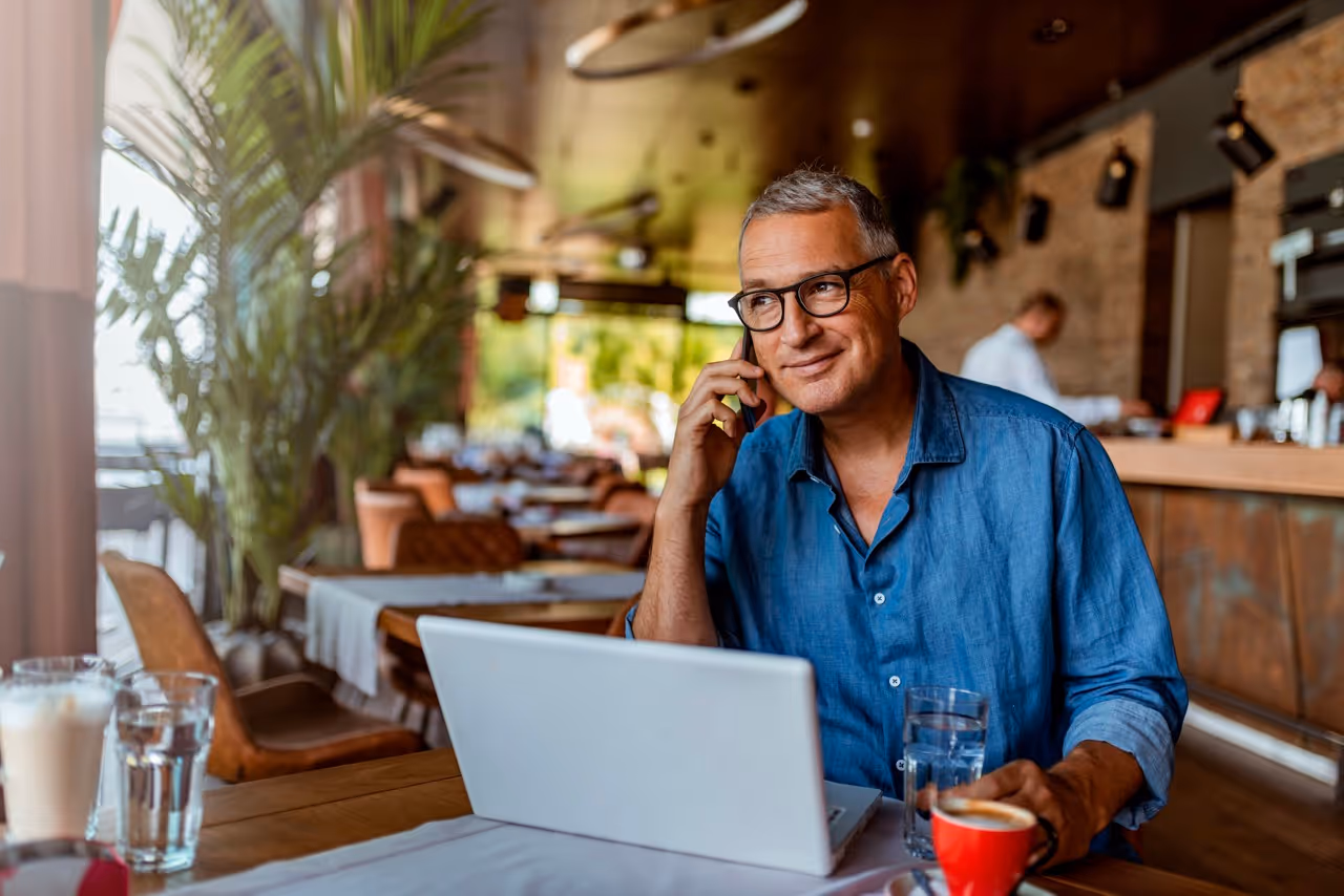 Middle-aged man in glasses using laptop and talking on phone at a cafe table with drinks.