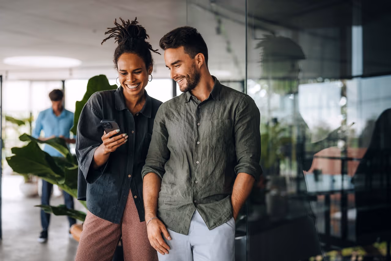 Two coworkers smile looking at a phone in a modern office with plants and glass walls.