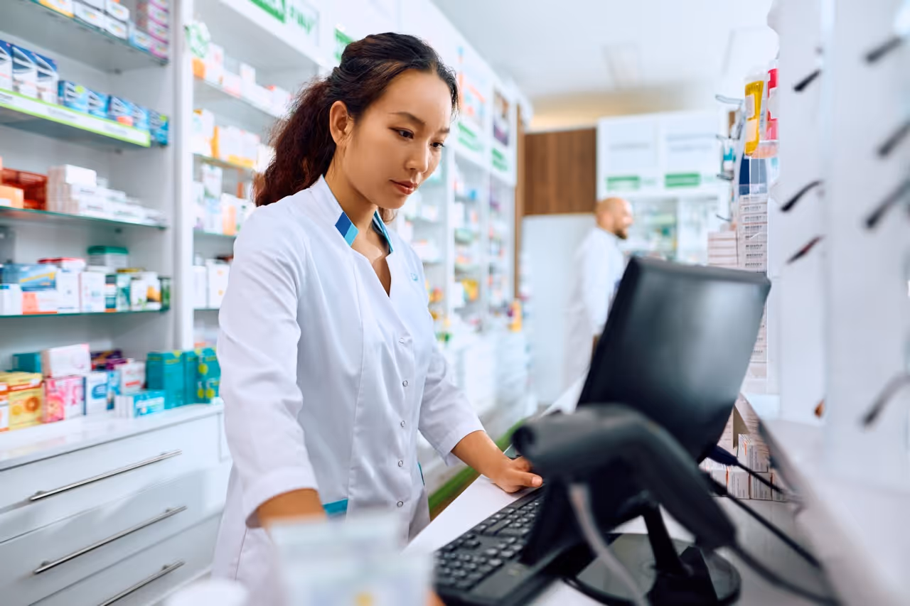 Pharmacist wearing white coat working at computer in a bright pharmacy with shelves of medicines.