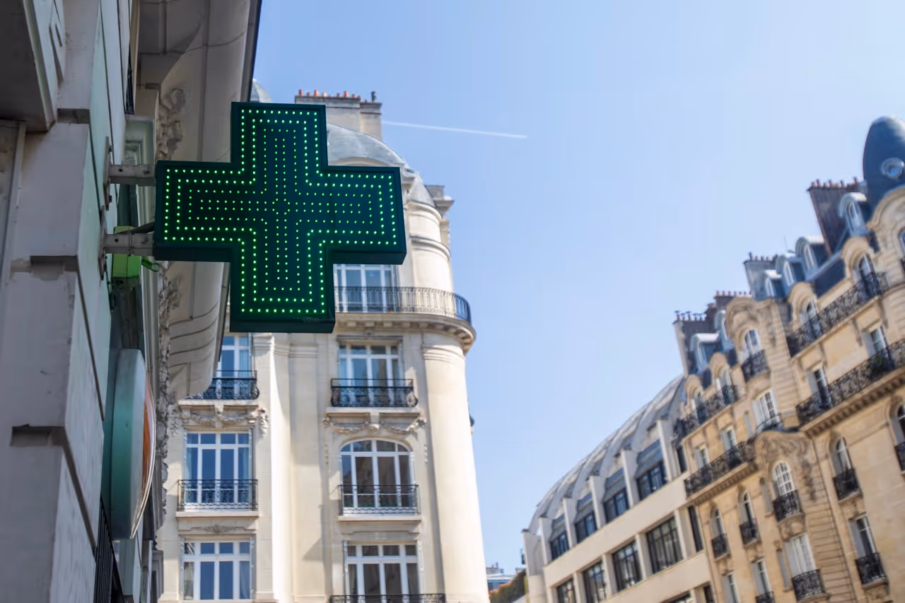 Green illuminated cross sign on building facade with Paris-style architecture in background under clear sky.