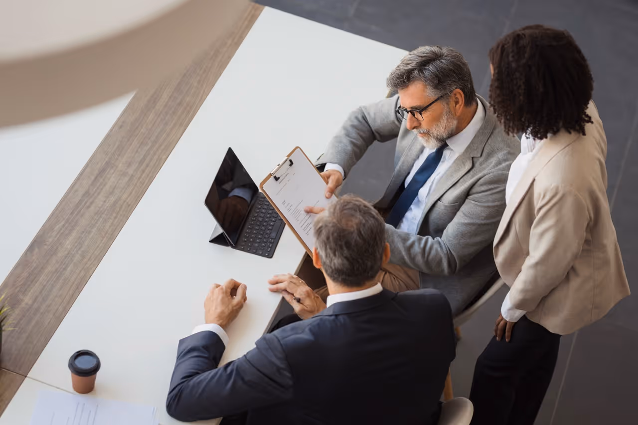 Three businesspeople reviewing a document on a clipboard at a white conference table with a tablet and coffee cup.