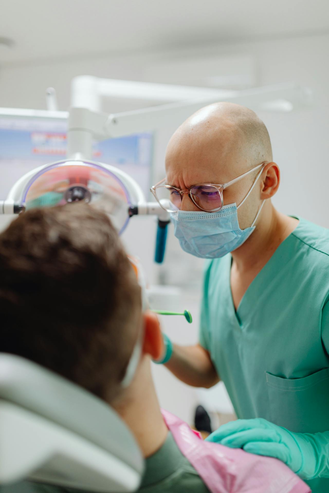 Dentist wearing glasses and mask examining a patient reclining in a dental chair.