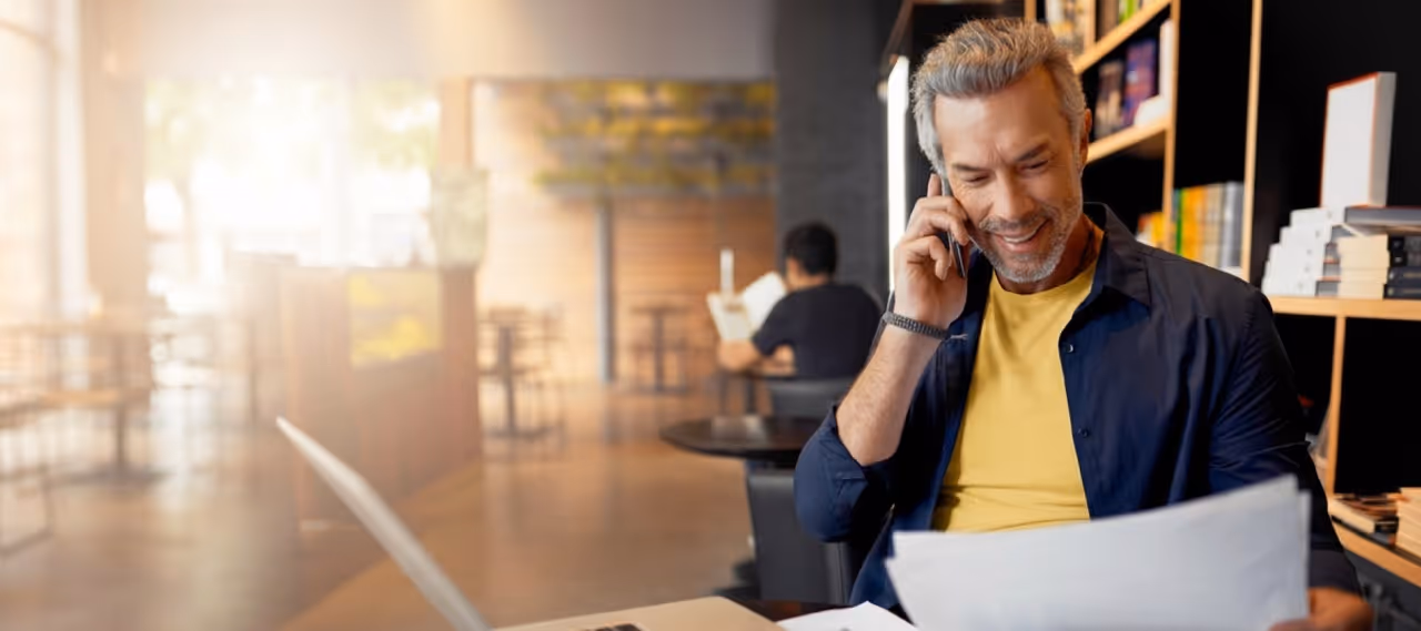 Smiling man talking on phone while reviewing documents in a cozy, well-lit cafe with bookshelves.