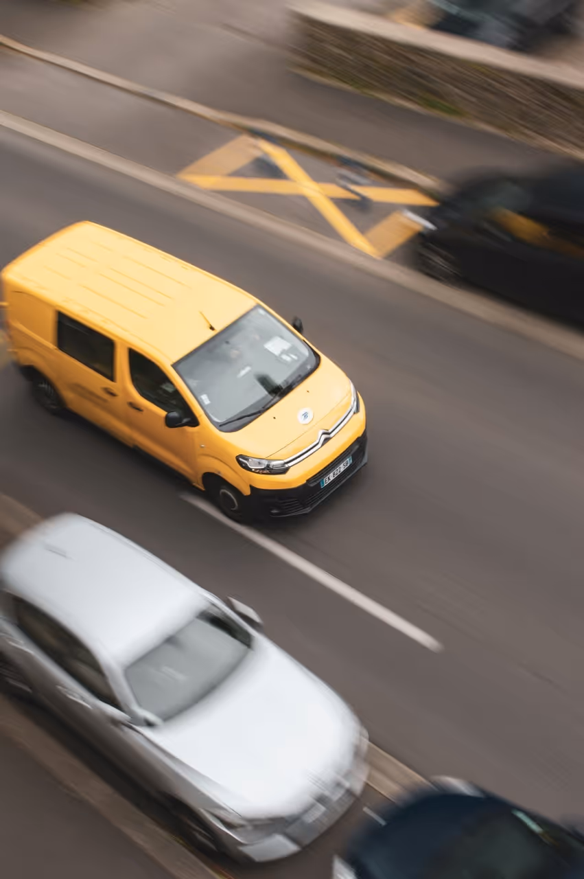 Yellow Citroën van driving on a city street with parked cars blurred nearby.