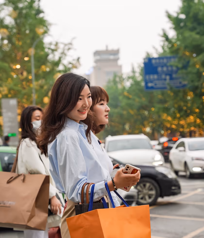 Two women holding shopping bags and phone, smiling on a busy city street.