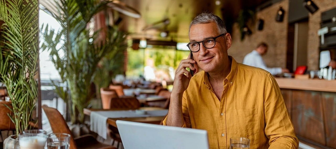 Man in a yellow shirt talks on phone while using a laptop in a cozy cafe setting.