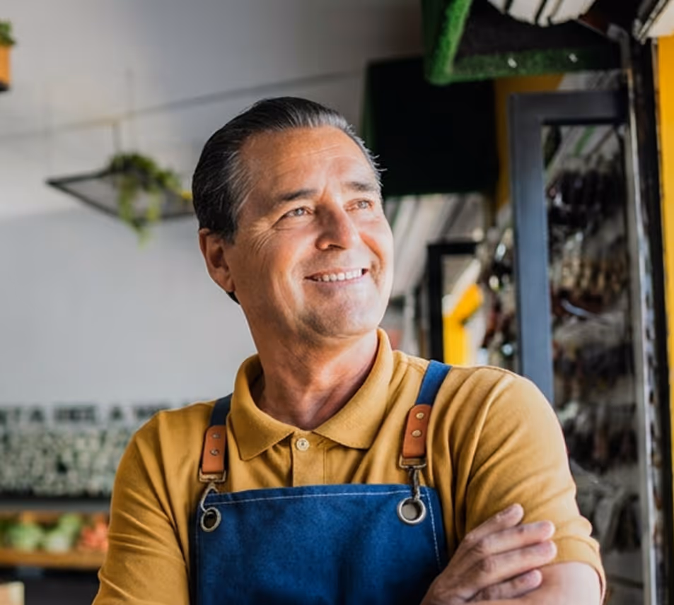 Smiling man wearing a mustard shirt and blue apron standing with arms crossed in a shop.