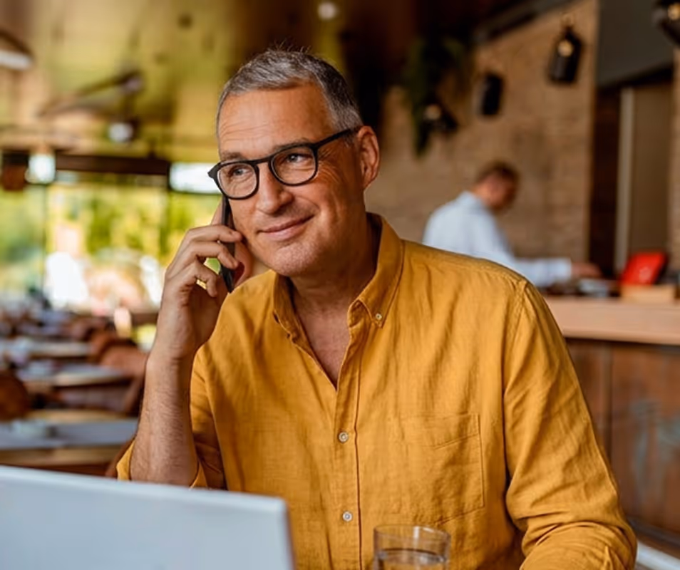 Man wearing glasses and a yellow shirt talking on phone inside a cafe with a laptop and glass of water.