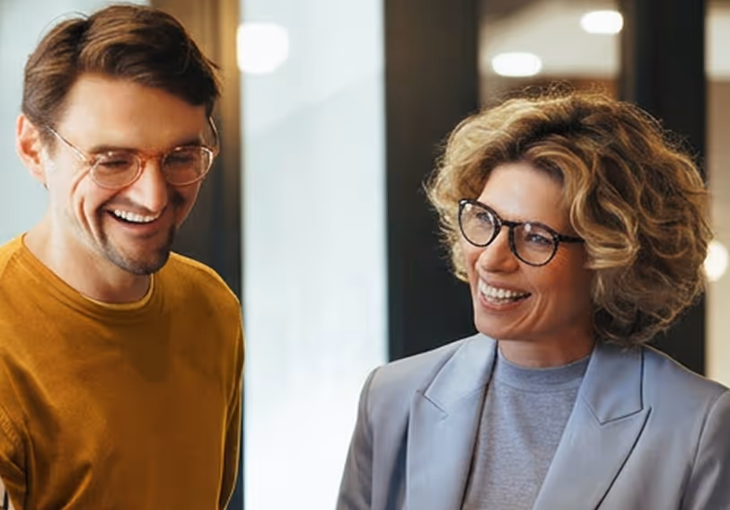 Man in mustard shirt and glasses smiling with woman in gray blazer and glasses in office.