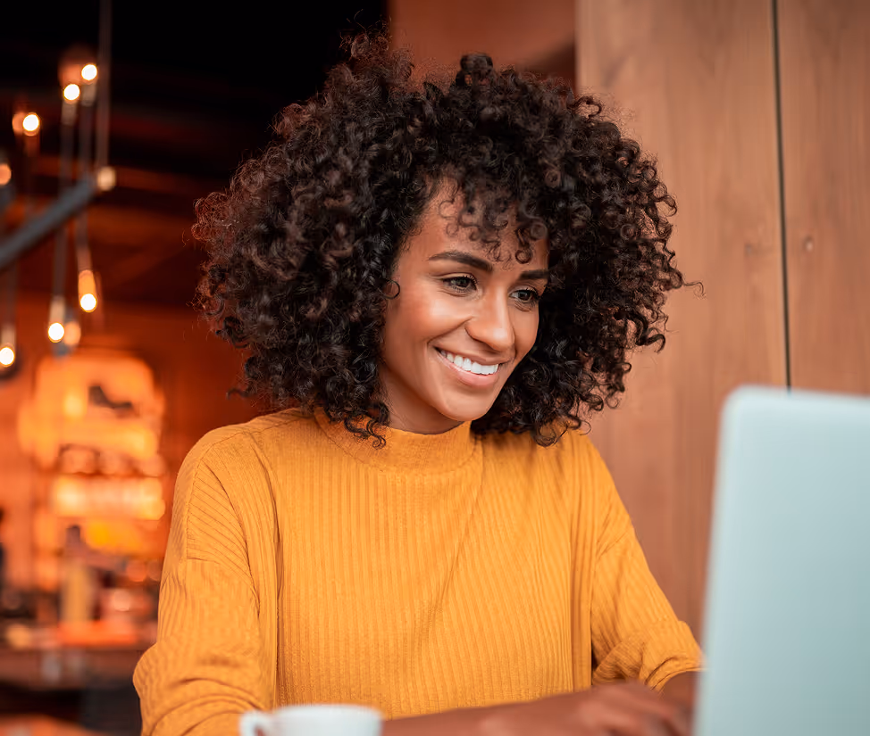 Smiling woman with curly hair in a mustard sweater working on a laptop in a cozy setting.