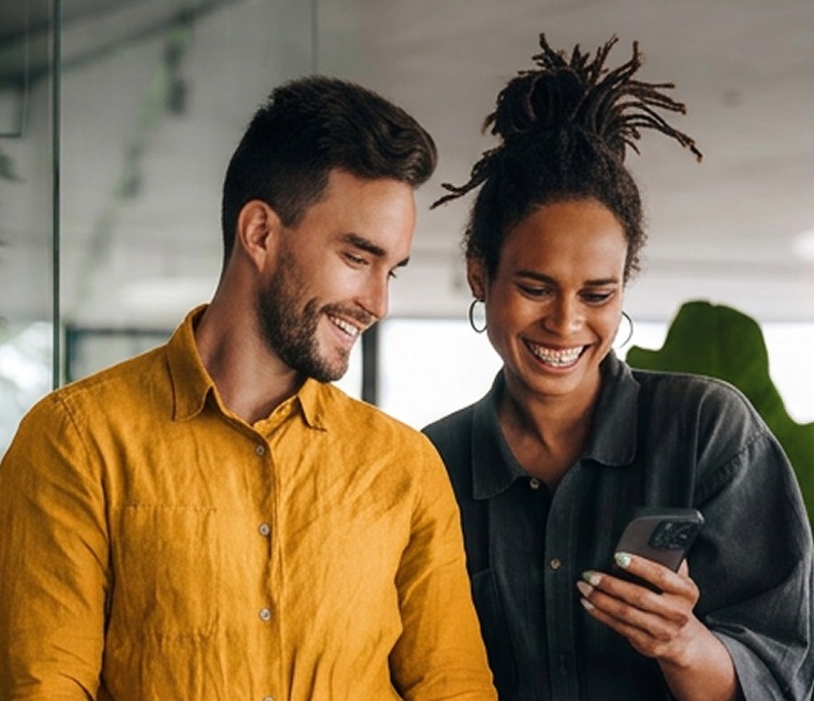 Man and woman smiling together as they look at a smartphone in a bright indoor space.
