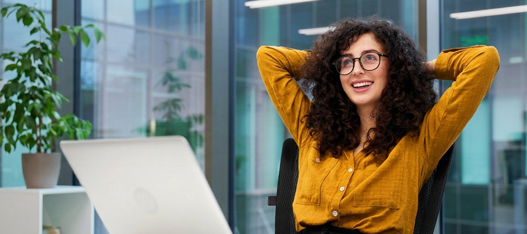 Smiling woman with curly hair and glasses wearing a mustard shirt, sitting relaxed by a laptop in an office.