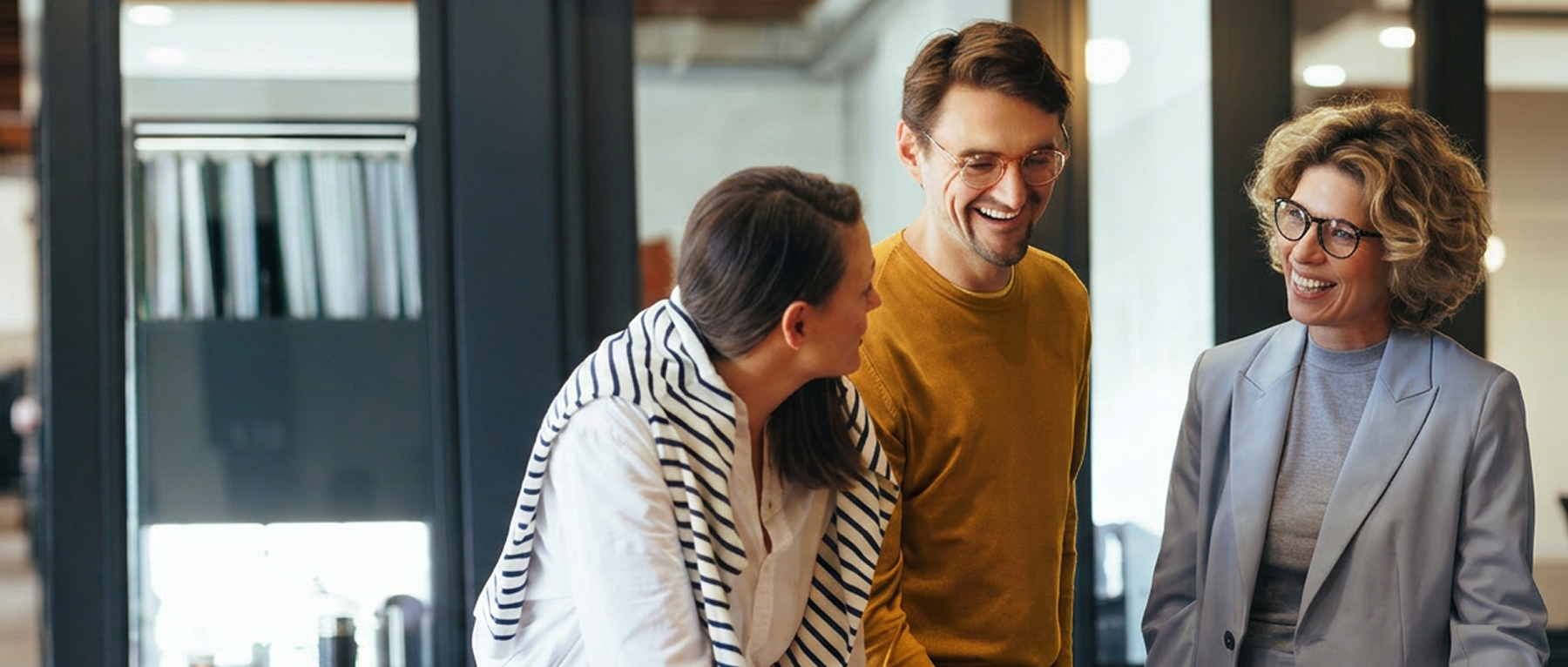 Three colleagues smiling and chatting in a modern office setting with glass walls.
