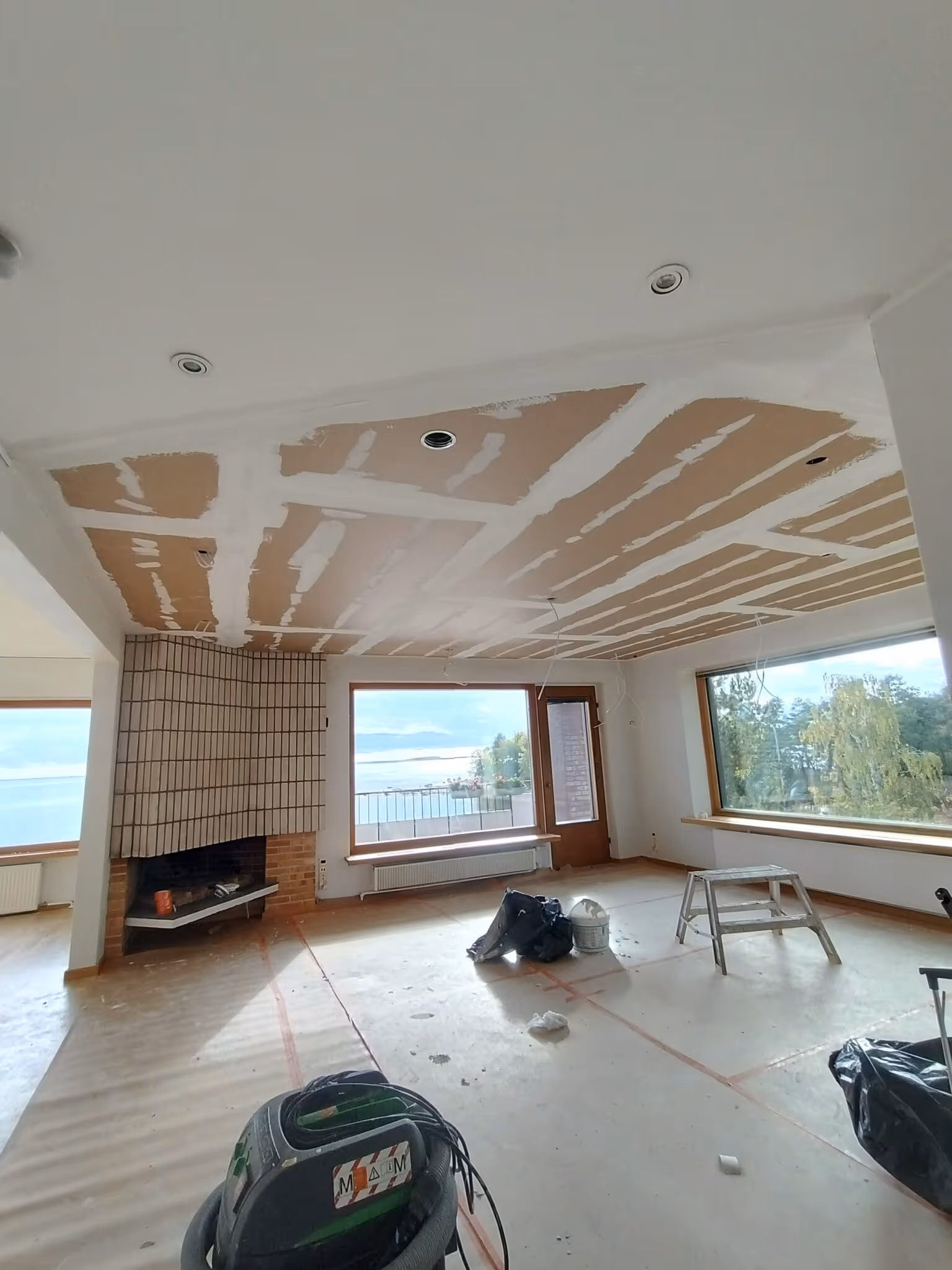 Huoneessa on katto, jossa on kipsilevyä ja valkoista maalia, sekä suuri tiilitakka. Ikkunoista näkyy merimaisema. Lattialla on suojamuovi ja työkaluja.