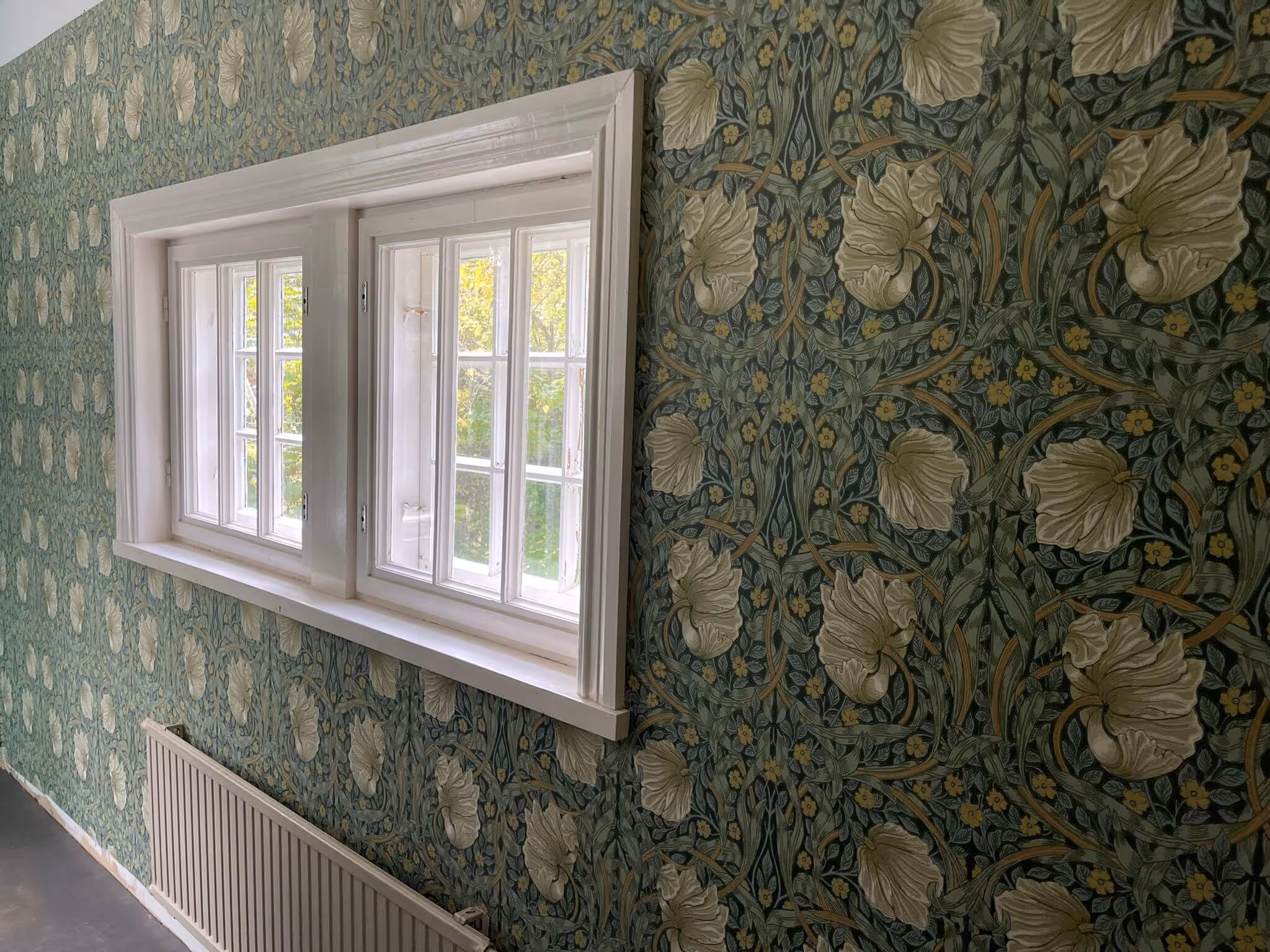 Huoneessa on ikkuna, jonka alla on patteri, ja seinät on tapetoitu kukka-aiheisella tapetilla.