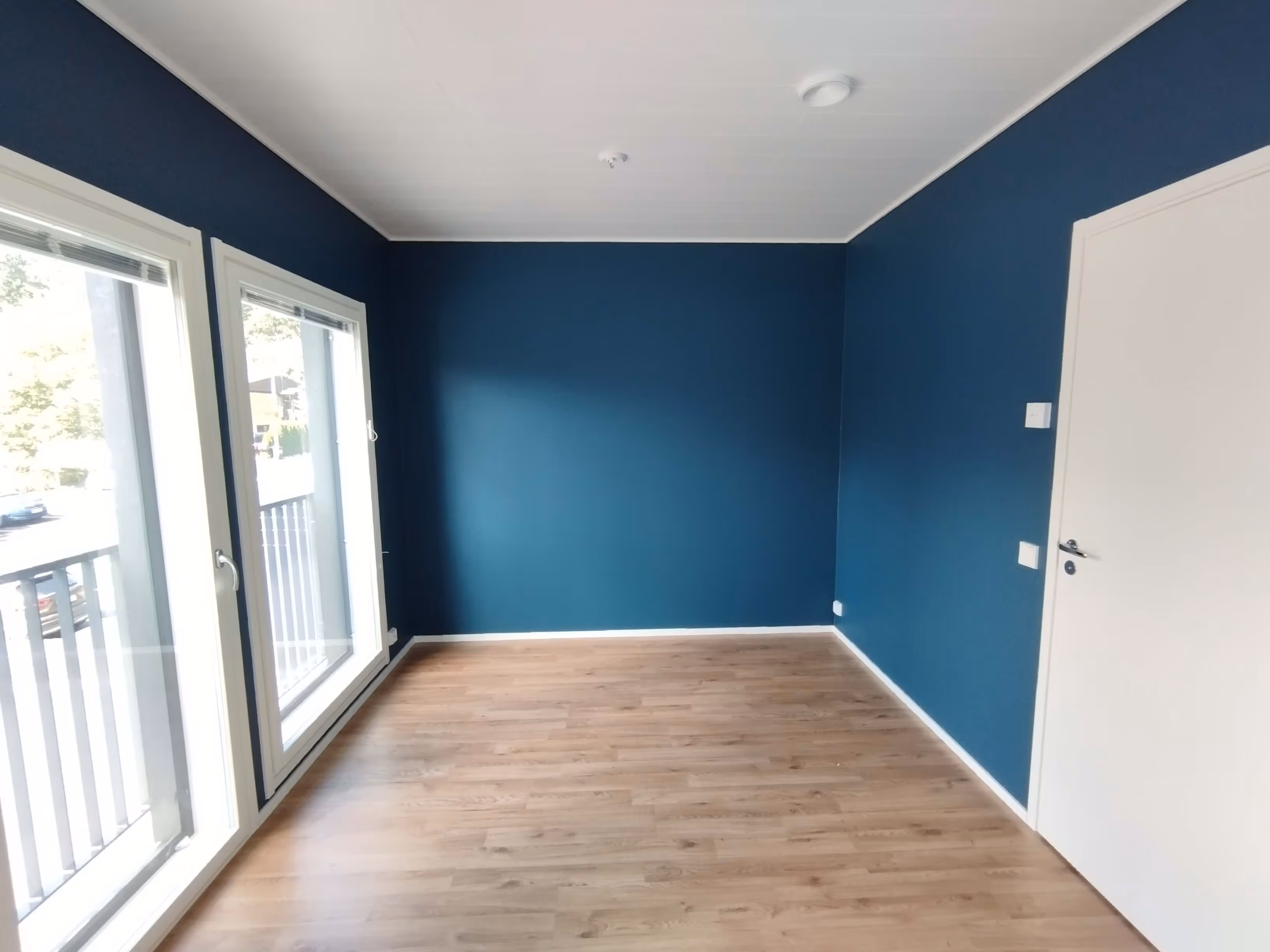 Huoneessa on siniset seinät, vaalea puulattia, valkoinen ovi ja kaksi ikkunaa, joista näkyy ulos.