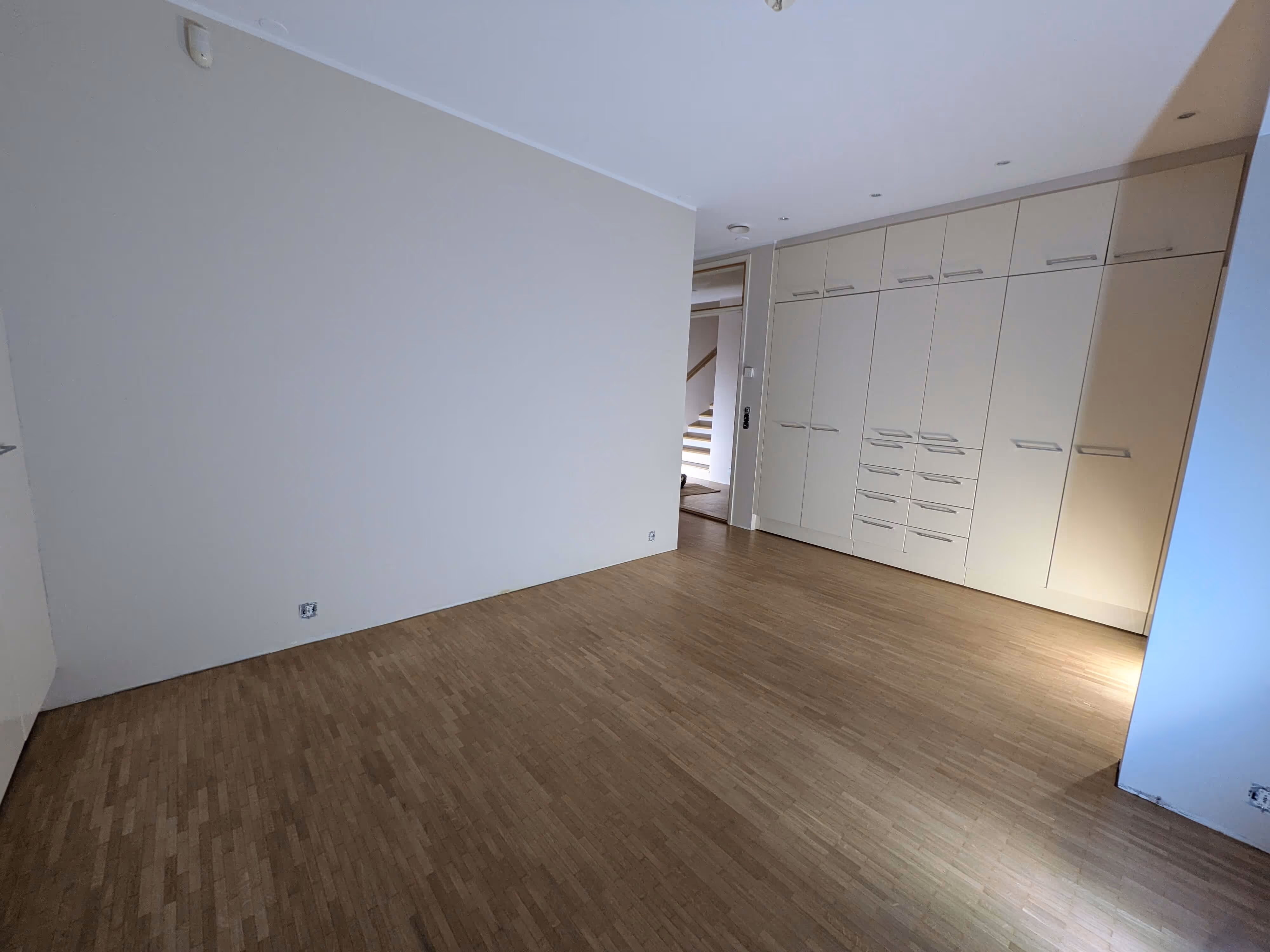 Tyhjä huone, jossa on vaaleat seinät, puulattia ja suuri valkoinen vaatekaappi. Portaiden näkymä avoimesta ovesta.