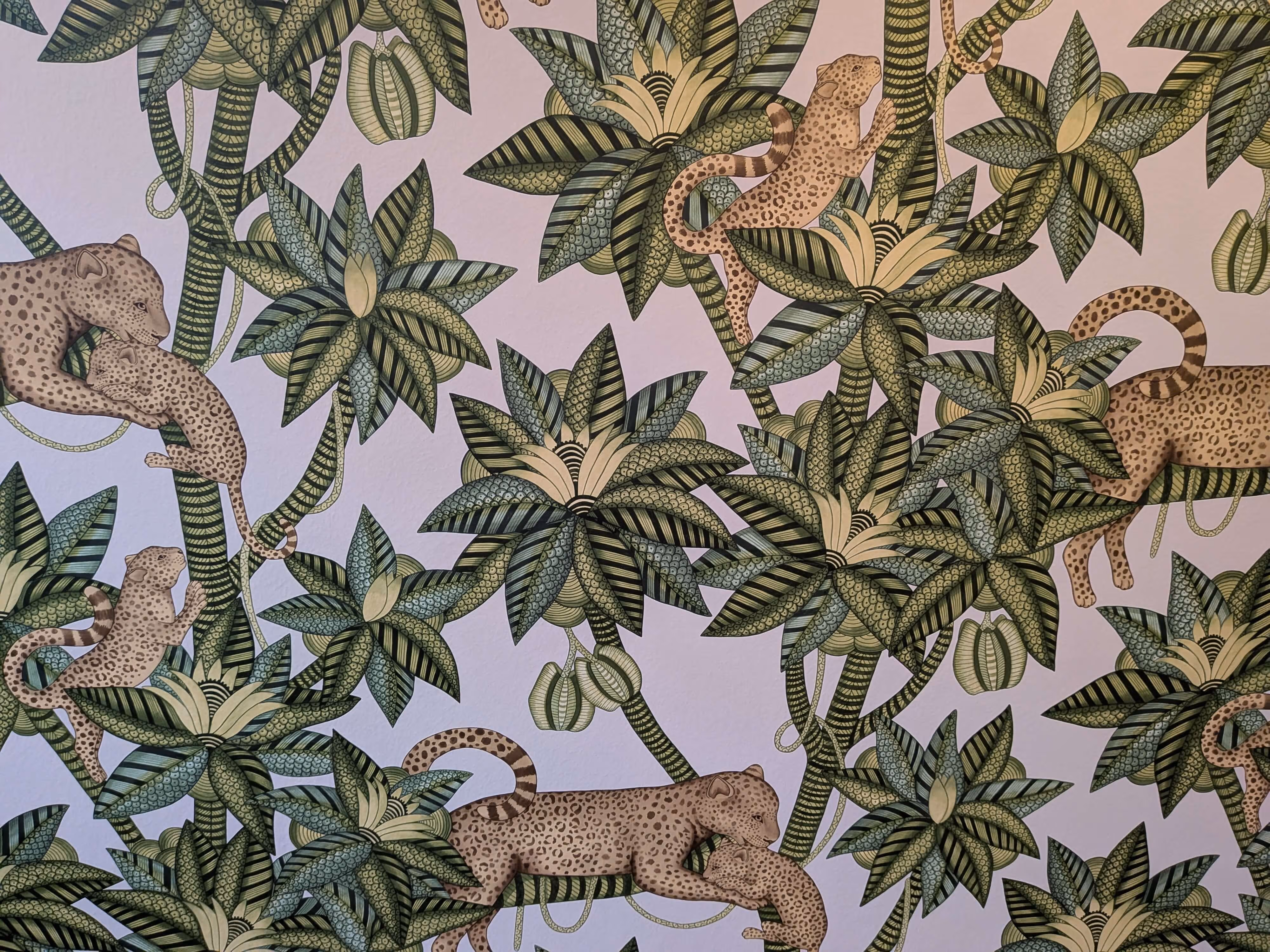 Kuvaus leikkisästä tapetista, jossa on leopardeja ja trooppisia kasveja vihreän, kerman ja ruskean sävyissä vaalealla taustalla.