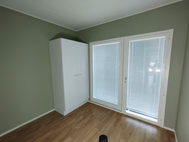 Huoneessa on valkoinen vaatekaappi, kaksi ikkunaa sälekaihtimilla ja puulattia. Seinät ovat vihreät ja valaistus on kirkas.