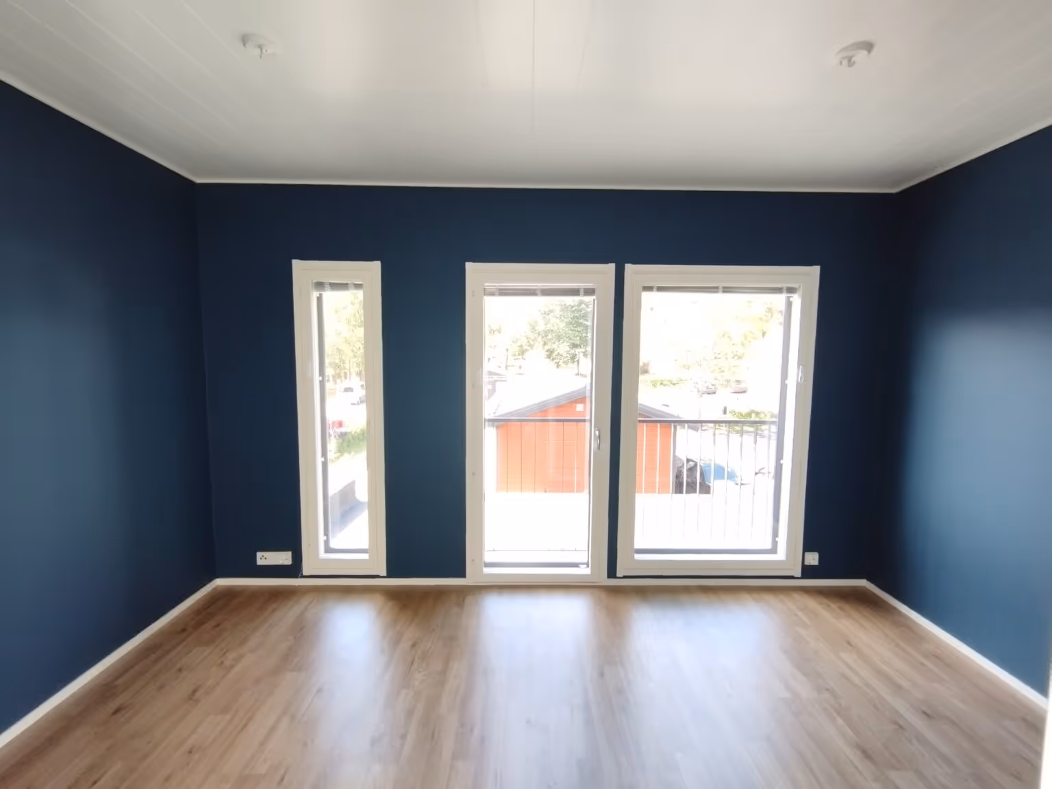 Huoneessa on tummansiniset seinät, vaaleat puulattiat ja kolme valkoista ikkunaa, joista näkyy ulos.