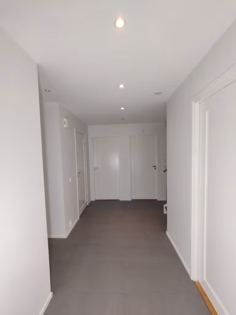Valkoinen käytävä, jossa on harmaa lattia, valkoiset seinät ja valkoiset ovet. Katossa on useita valaisimia.
