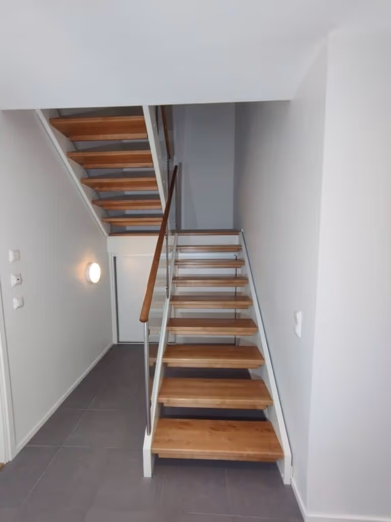 Sisätiloissa on puuportaat, joissa on valkoiset sivut ja metallinen kaide, sekä harmaat lattialaatat ja valkoiset seinät.